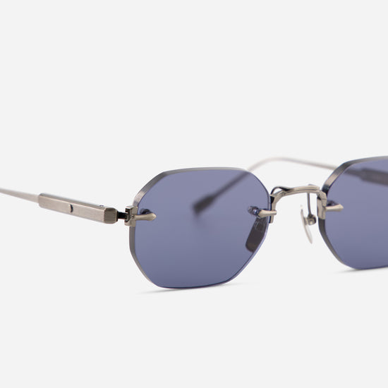 Close-up of rimless sunglasses with blue-gray lenses and a antique silver titanium frame, shown against a light gray background.
