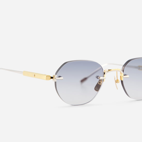 Close-up of a pair of rimless sunglasses with gradient gray lenses, gold metal bridge and temple accents, and white temples on a light gray background.